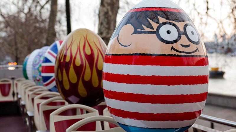 Six eggs on an open top bus, with Where's Wally at the front, a gold egg with red fire-like painting behind, and behind that is the Union Jack. 