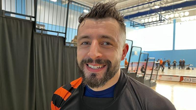 A man with spikey hair and a brown beard in a black and orange strip smiles at the camera. 