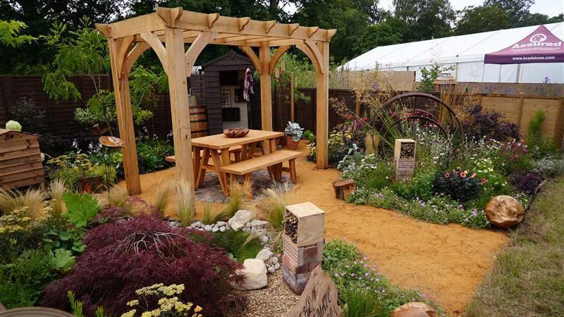 A garden at a garden show. There is a wooden pergola with two benches either side of a wooden table underneath. Planting can be seen either side with an insect hotel and a wheel shaped sculpture.