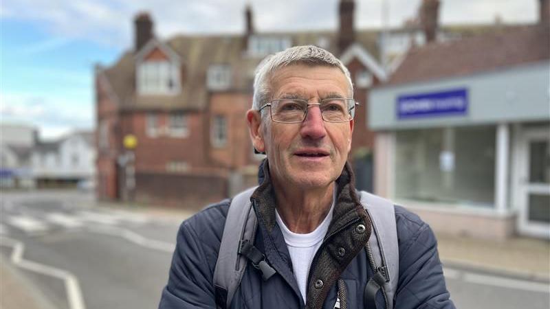 A man in a black jacket on an English High Street. He has grey hair and glasses.