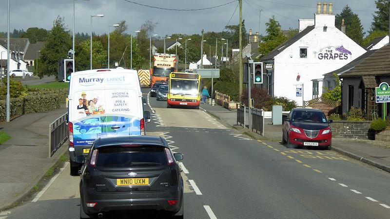 Multiple cars, a van, a bus and a lorry travelling through the centre Springholm village on the A75. A restaurant and housing is shown on the side of the road and a set of traffic lights are positioned half way up the road.