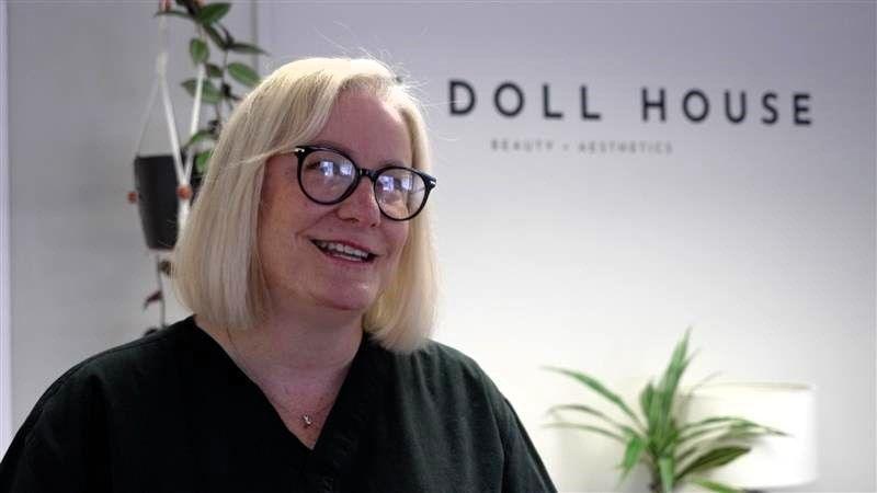 Emma Brown in her beauty salon. The DOLL HOUSE logo is behind her on a wall. A couple of plants are in front of the wall.