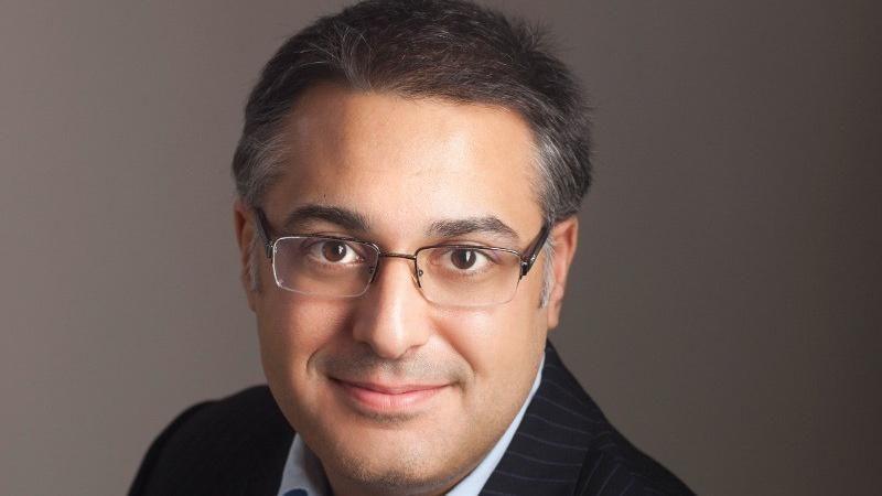 A headshot of surgeon Yaser Jabbar, wearing glasses and a dark suit, with dark grey hair