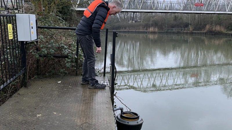 Small circular bin rests just above the River Nene. A man in a life jacket stands on a metal platfrom next to the river looking down at the seabin. 