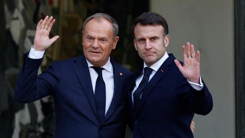 Donald Tusk and Emmanuel Macron, dressed in suits, wave as they are photographed standing next to each other