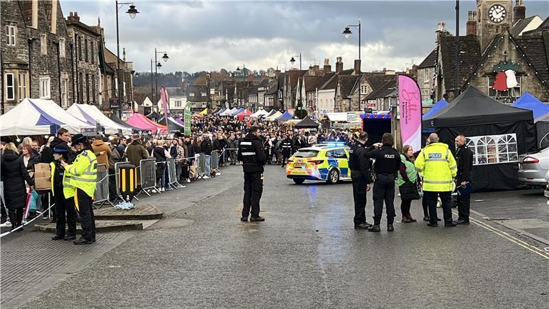 A picture of the high street in Chipping Sodbury with barriers set up on the left side of the road. Behind the barriers there are lots of people, stretching all the way down the road into the distance. There is a police car parked in the middle of the road, and several police officers speaking to bystanders. 