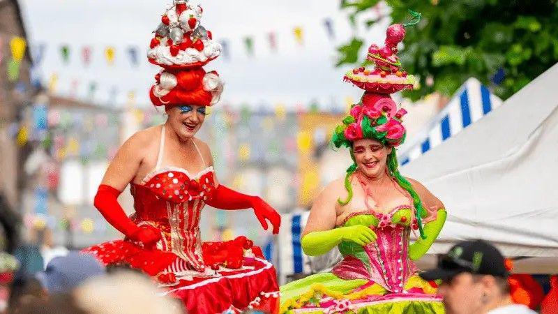 Two people are dressed up on stilts with a market in the background. One has a red wig with a huge pink and red hat in the style of a cake. They are wearing red gloves, blue sparkly eyeshadow and a halter neck red and white puffy dress, The other person has a pink and red cake-style hat, with a green wig. They are wearing a pink and green large dress that billows out.
