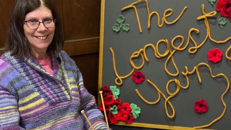 Claire Holmes from St Macartins' Knitwits wearing a purple, yellow and blue knitted jumper is pictured smiling beside a sign reading 'The Longest Yarn'.