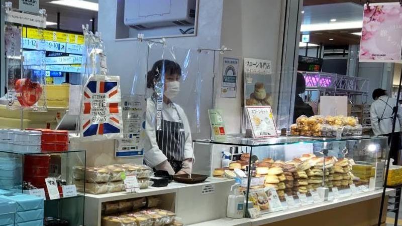 Under the bright lights of a railway station in Japan, there are lots of scones stacked up with prices and descriptions in Japanese and a server waiting