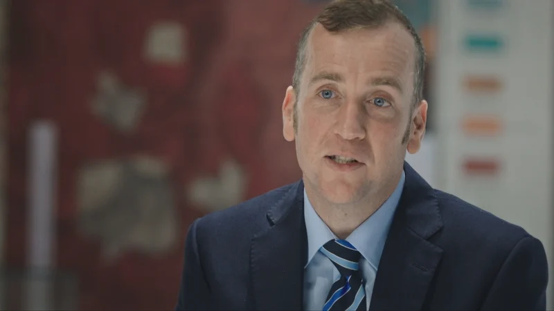 Simon O'Donovan. He has short brown hair, blue eyes, wearing a navy blazer, light blue shirt and blue striped tie.