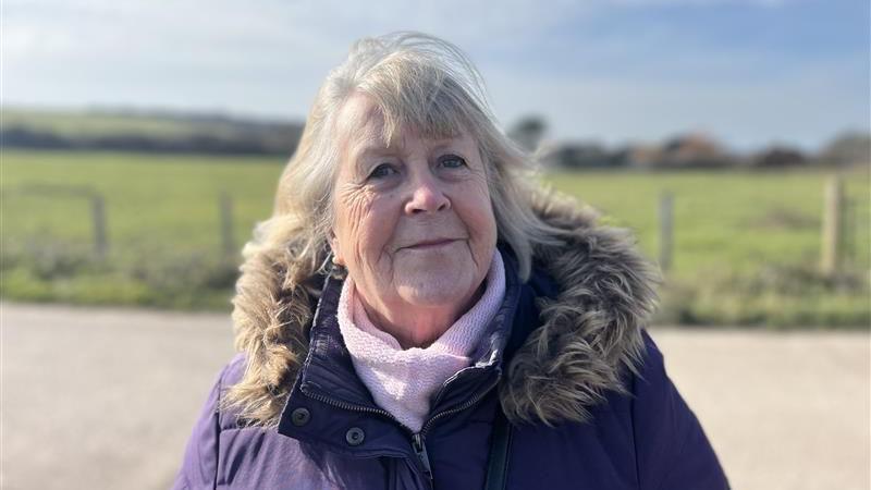 A woman with white hair in a blue parka coat stands in front of a green field.