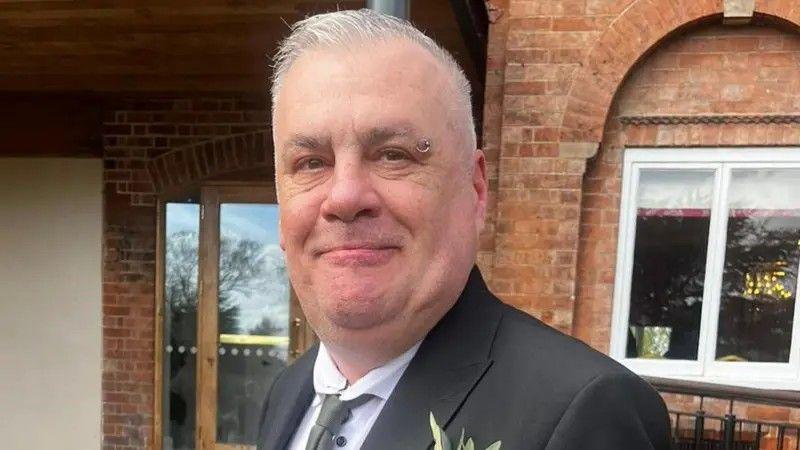 A man with short cropped grey hair and a ring piercing on the end of his left eyebrow. He is wearing a black blazer over a white shirt and a grey tie. There is a flower in the right breast pocket. He is standing in front of a red-bricked building and smiling.