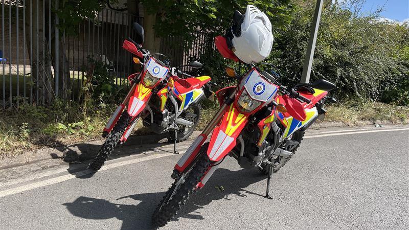 Two red police off-road bikes propped up side by side on the edge of a road. A white helmet is hanging from the handlebars of one of them. Behind the bikes is a hedgerow, and beyond that there's blue sky. The bikes are mostly red, with yellow and blue patches on the mudguards. On the front is a West Midlands Police badge. 