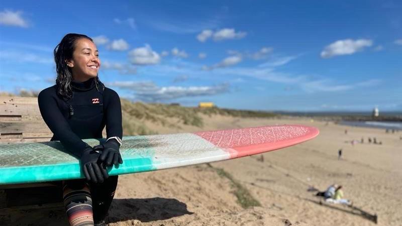 Stephanie Smailes sits on Coney beach in a winter wetsuit with wetsuit gloves on. She is holding a pink, white and turquoise board resting on her lap. She has dark long hair and is smiling and looking to the right of the camera out to sea.