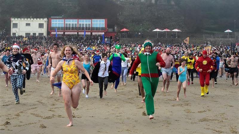 Crowds of people dressed as superheroes run towards the camera grinning down the beach. 