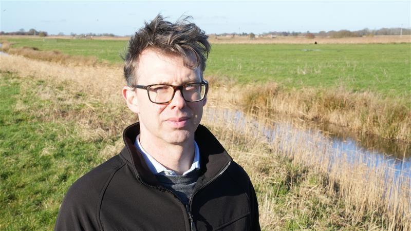 Eliot Lyne is looking at the camera and is stood in a field. He is wearing glasses and is wearing a black sweatshirt, shirt and grey jumper. 