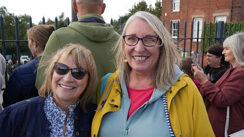 A woman with a light coloured bob wearing sunglasses, a blue coat and a blue blouse stands next to a taller woman with grey/blonde hair, wearing a yellow raincoat, a light blue jumper and a red top. They are both smiling and people are standing behind them.