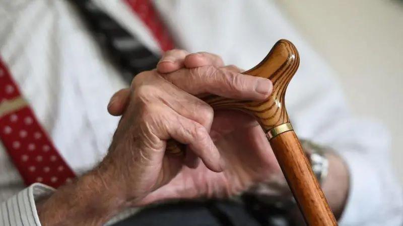 A close of an elderly man's hands. He is holding a walking stick and wearing a white shirt and a tie. 