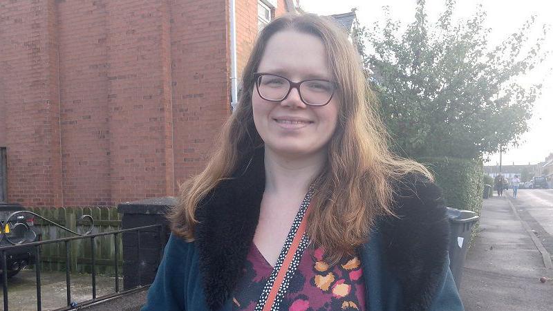 Catherine Coffey  smiling at the camera. She has long ginger hair, wearing brown framed glasses, a velvet suede furred coat and a purple, orange and black floral patterned top. She is standing on a pavement with bins behind her.