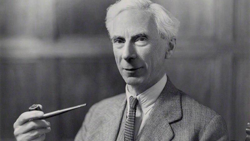 A black and white picture of Bertrand Russell looking at the camera. He has short white hair and is wearing a shirt, a tie and a jacket. He is holding a pipe in one hand.