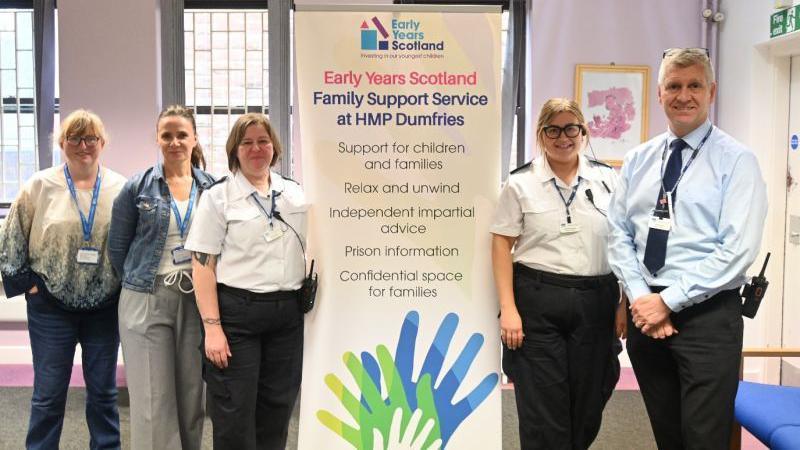 Four adults stand around a sign describing the Early Years Scotland Family Support Service at HMP Dumfries.