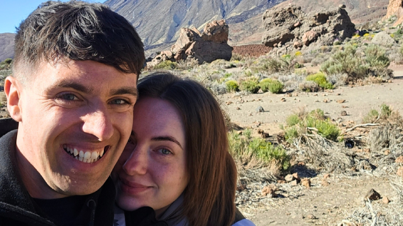 Jack Cope and Chloe are taking a selfie of them together in a mountain terrain. Jack, on the left, has short brown hair and is wearing a black jacket. Chloe, on the right, has long brown hair and is wearing a lilac jacket.