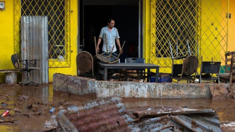 A man stands in the doorway of a bright yellow building, in front of him the ground is completely  covered in mud with twisted corrugated metal, broken furniture and scattered rubbish. Tables and chairs are also piled up and splattered in mud.