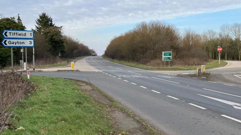 A road junction showing Tiffield, and Gayton, to the left. It shows a road that veers to the right, a dual carriageway that has a lorry on it in the distance, another junction to the right, with no entry signs, and another slip road. There are trees, bushes and grass around the junction. 