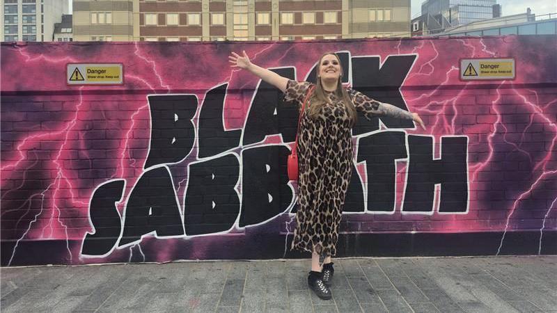 Kirsty Bosley pictured with her arms spread wide and smiling. She is stood against the black and pink Black sabbath wall mural in a cheetah print jumpsuit with chest length hair.