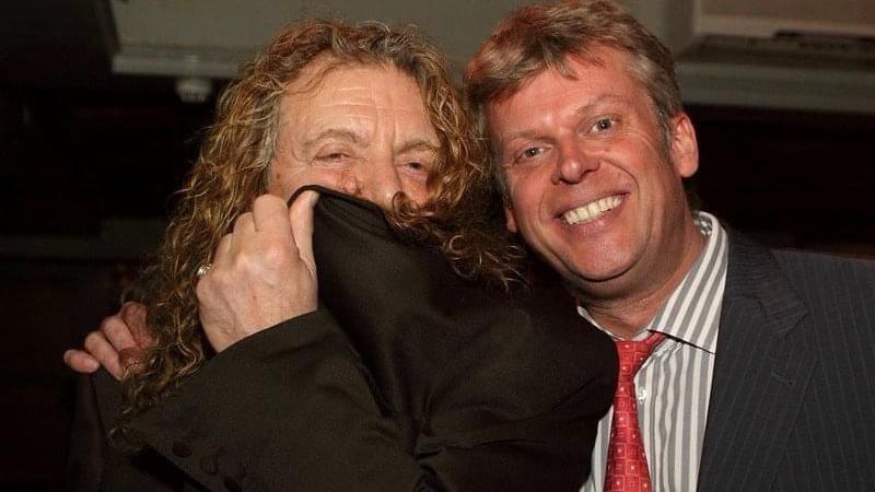 Robert Plant, who has long curly hair, partially covers his face with a black coat and stands next to another man, John Bunce, who has short brown hair. is smiling and wearing a pinstripe suit with a striped shirt and a red tie.