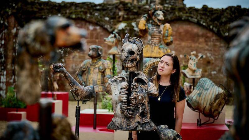 A woman with dark hair and a dark top looking at a statue of a skeletal figure in an area surrounded by other statues