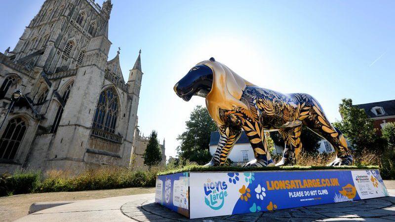 A lion sculpture painted in black and gold is stood in front of Gloucester cathedral. The sun is shining and the sky is blue.