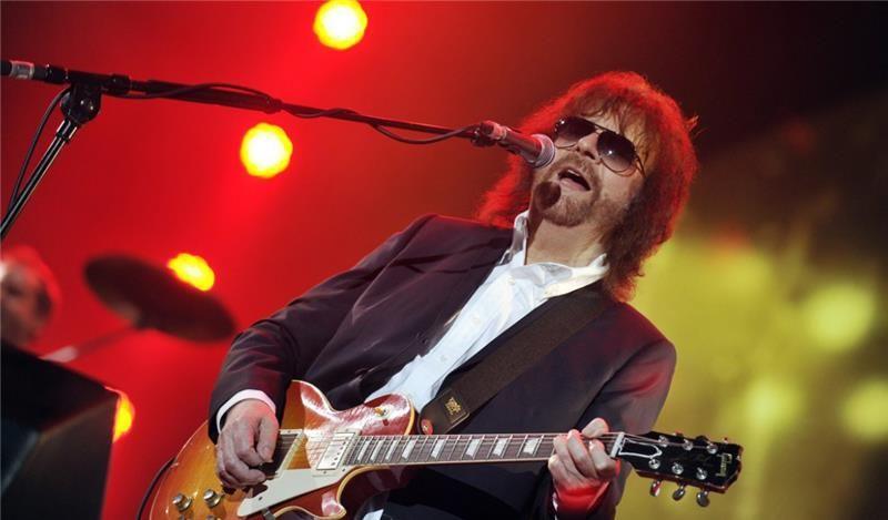 Jeff Lynne plays a guitar and sings into a microphone on stage. He wears a purple suit jacket and a white shirt