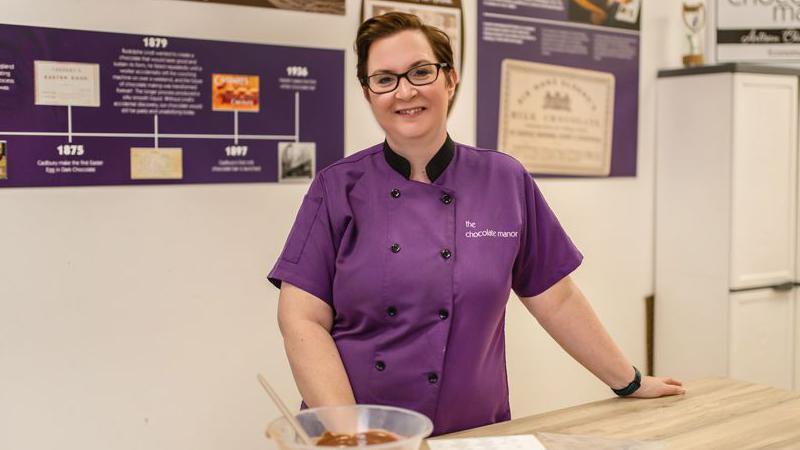 Geri Martin - a woman wearing a purple chef coat with 'The Chocolate Manor' in white embroidery. She is standing at a wooden kitchen island with a bowl of chocolate in front of her.