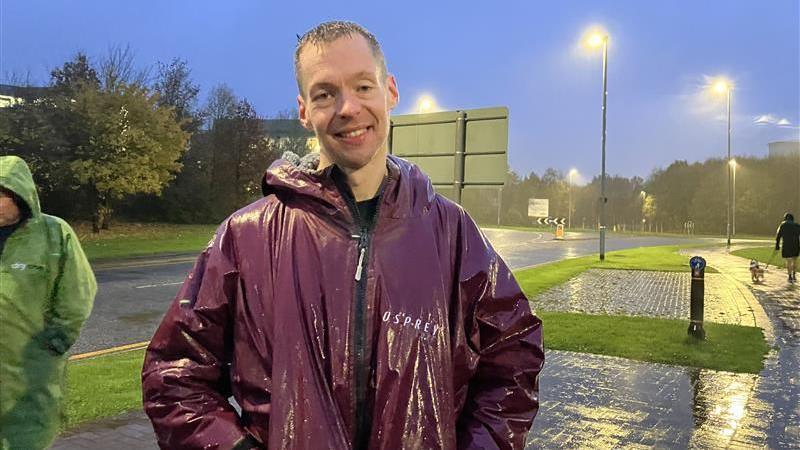 Group leader Dan Winfield wearing a maroon coat, smiling for the camera. He is wet from the rain. A person is seen walking a dog behind him.