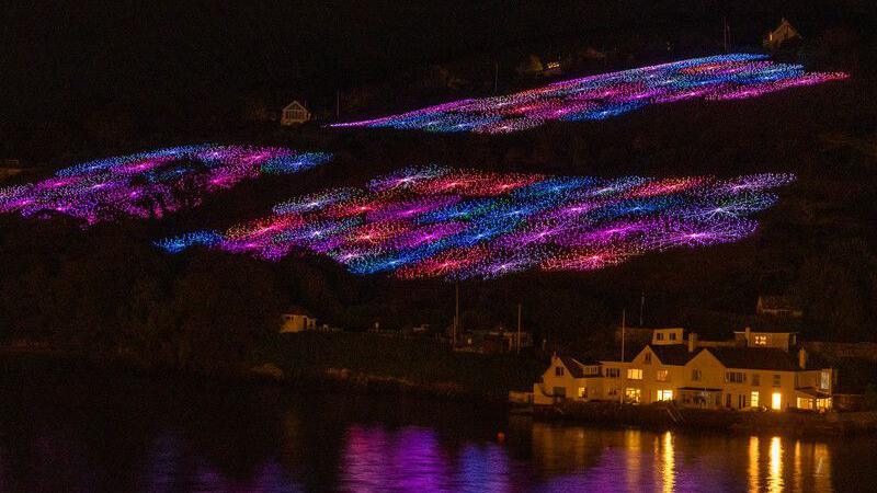 Patches of fibre optic lights light up the hillside at night. They spiral out in pink, purple, blue, orange and white patches. There is a house or hotel in the bottom right of the image which has its lights on.