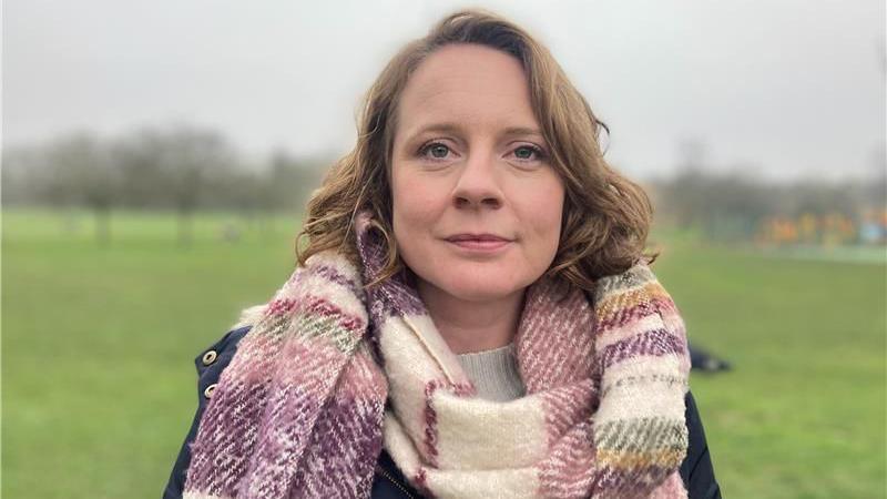 Cheney Payne in a park. She wears a large pink and cream checked woollen scarf and dark coat. 