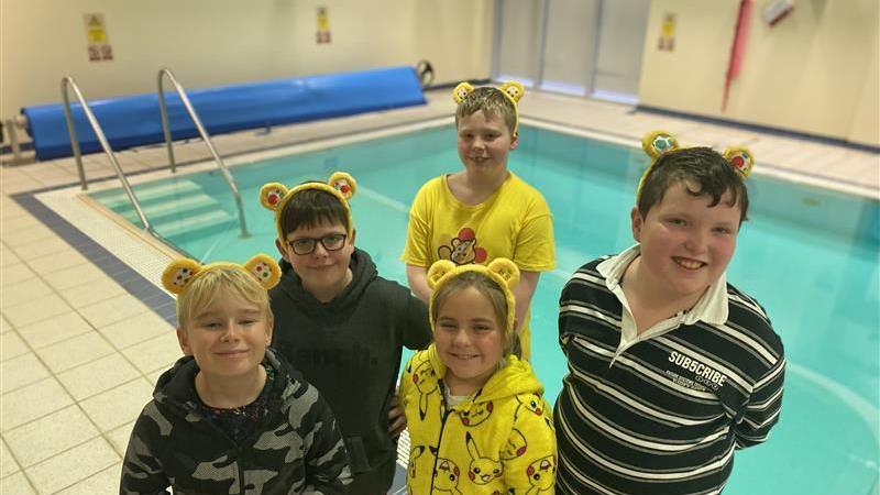 Five pupils from Le Rondin school stood in front of a swimming pool. four boys and a girl in the centre. They are all wearing a pair of yellow Pudsey ears