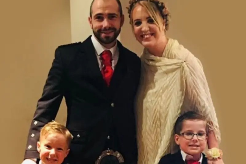 Barry Shelley, and their two children at a wedding - Barry is dressed in a suit with kilt, Shelley is in a white dress and both boys are dressed formally, with black jackets, white shirts and red ties.