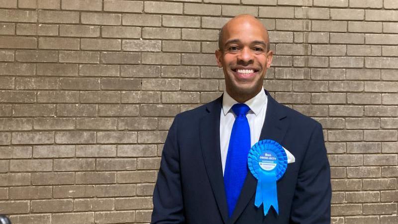 Ben Obese-Jecty  smiling and wearing a blue Conservative rosette