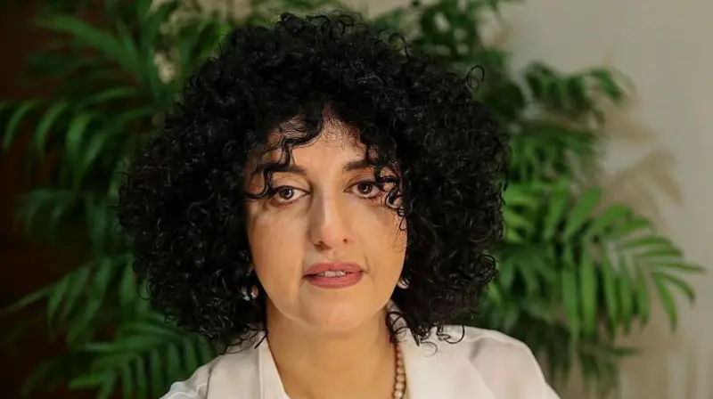 Portrait of a woman with voluminous curly black hair wearing a white blazer and beaded necklace, looking directly at camera against a soft background with green plants