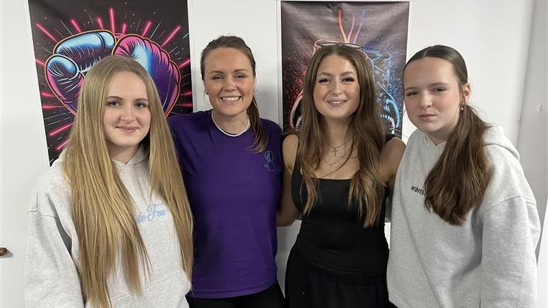 Four women standing together, all looking into the camera and smiling. All have long hair, two wearing hoodies, one a T-shirt, another a dress.