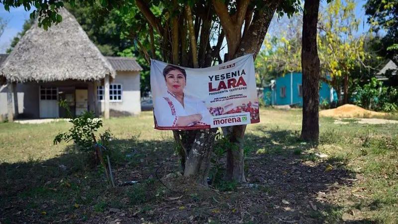A poster for Yesenia Lara who ran for mayor is hung on a tree in front of a thatched and tiled house