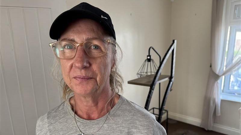 A woman with blonde hair tied back and glasses. She is wearing a grey tshirt and a black cap.