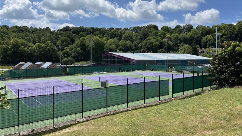 Three outdoor tennis courts at Withdean, which are purple with a green outside perimeter. They are surrounded by grass and trees. It is a sunny day.