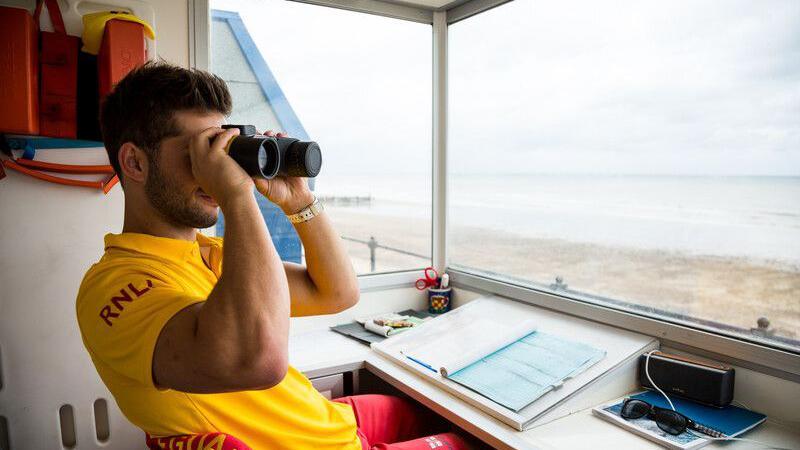 A lifeguard sits in a lifeguard hut on a beach. He looks into binoculars and monitors the beach. He wears a bright yellow t shirt with the letters RNLI written on his arm. He has short brown hair and wears a watch of his left wrist.  