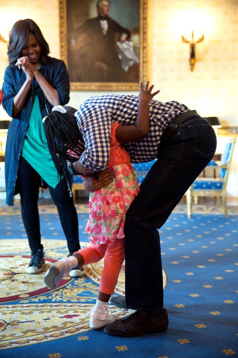 The First Lady watches as President Obama gives a hug to a little girl in the White House. the President is bent down to reach the small child.