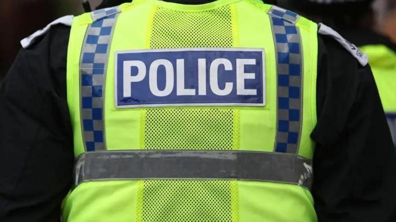A stock image of the back of a police officer. They are wearing a black jacket and yellow hi-vis vest over the top that has 'POLICE' written on it in white capital letters, surrounded by a blue box.