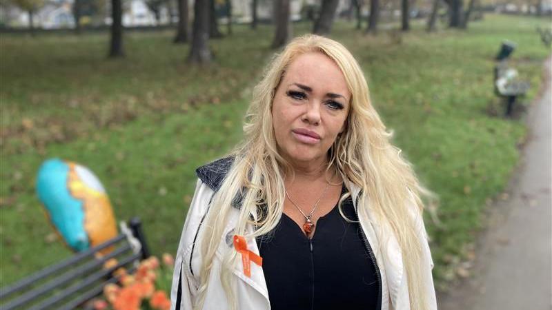 A woman, who wears an orange ribbon, stands next to a memorial bench in a park. Orange roses lay on the bench and a Garfield balloon is tied to it.