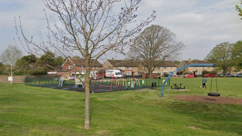 A small play area in a green field with eight houses in the background. There is a small brown tree in the foreground